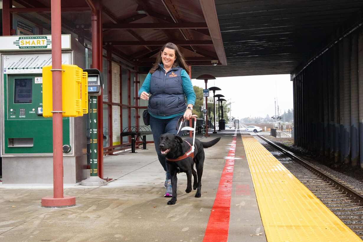 A guide dog mobility instructor works with a dog on a train station platform.