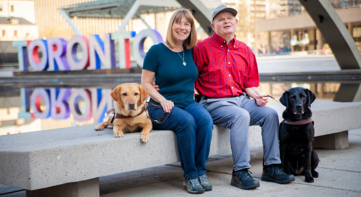 A married couple sit on a bench with a large "Toronto" sign in the background; the people both have guide dogs at their sides.