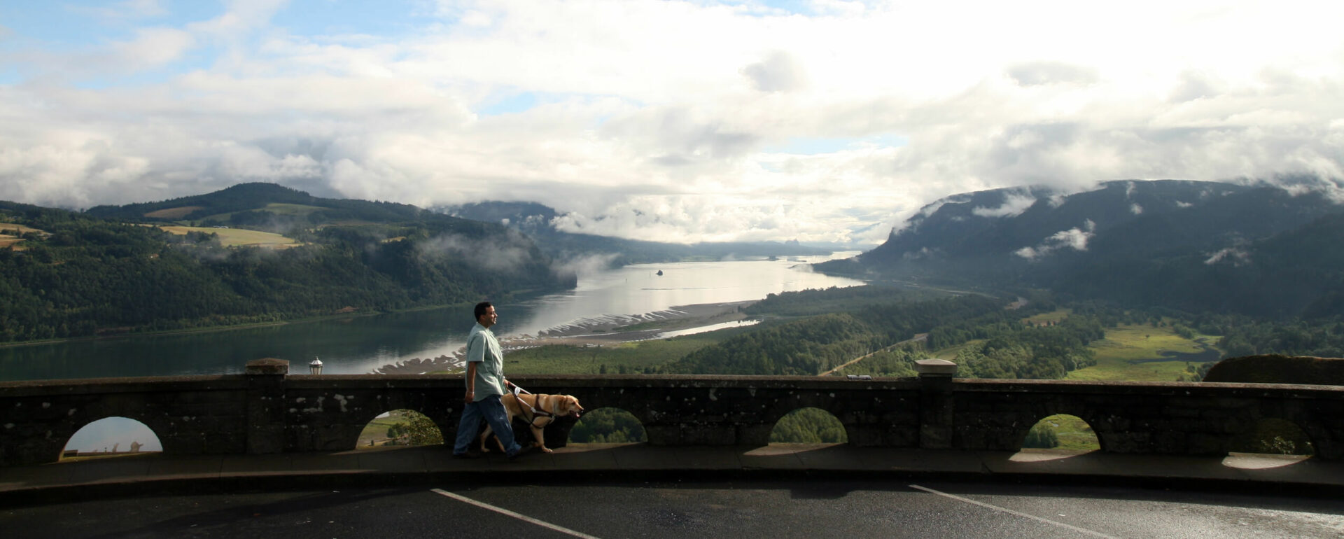 A GDB graduate walks with his yellow Lab guide dog along Crowne Point in Oregon with the beautiful Columbia Gorge in the background