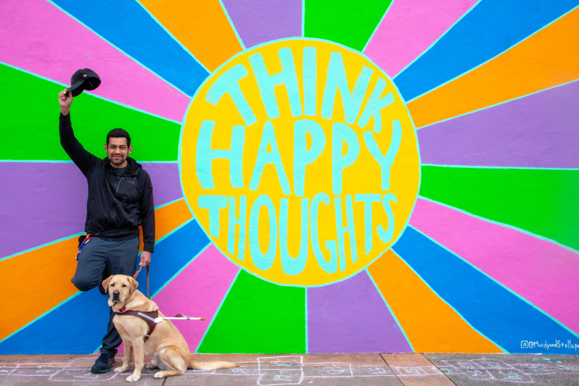 A man with a guide dog stands in front of a colorful sunburst mural that reads "Think Happy Thoughts."