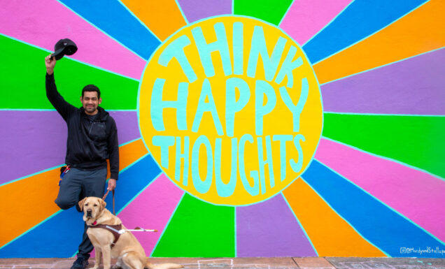 A man with a guide dog stands in front of a colorful sunburst mural that reads "Think Happy Thoughts."