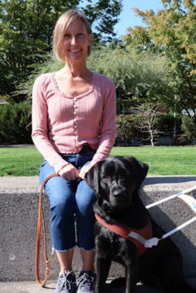 A woman sits on a low wall with a black Lab guide dog by her side.