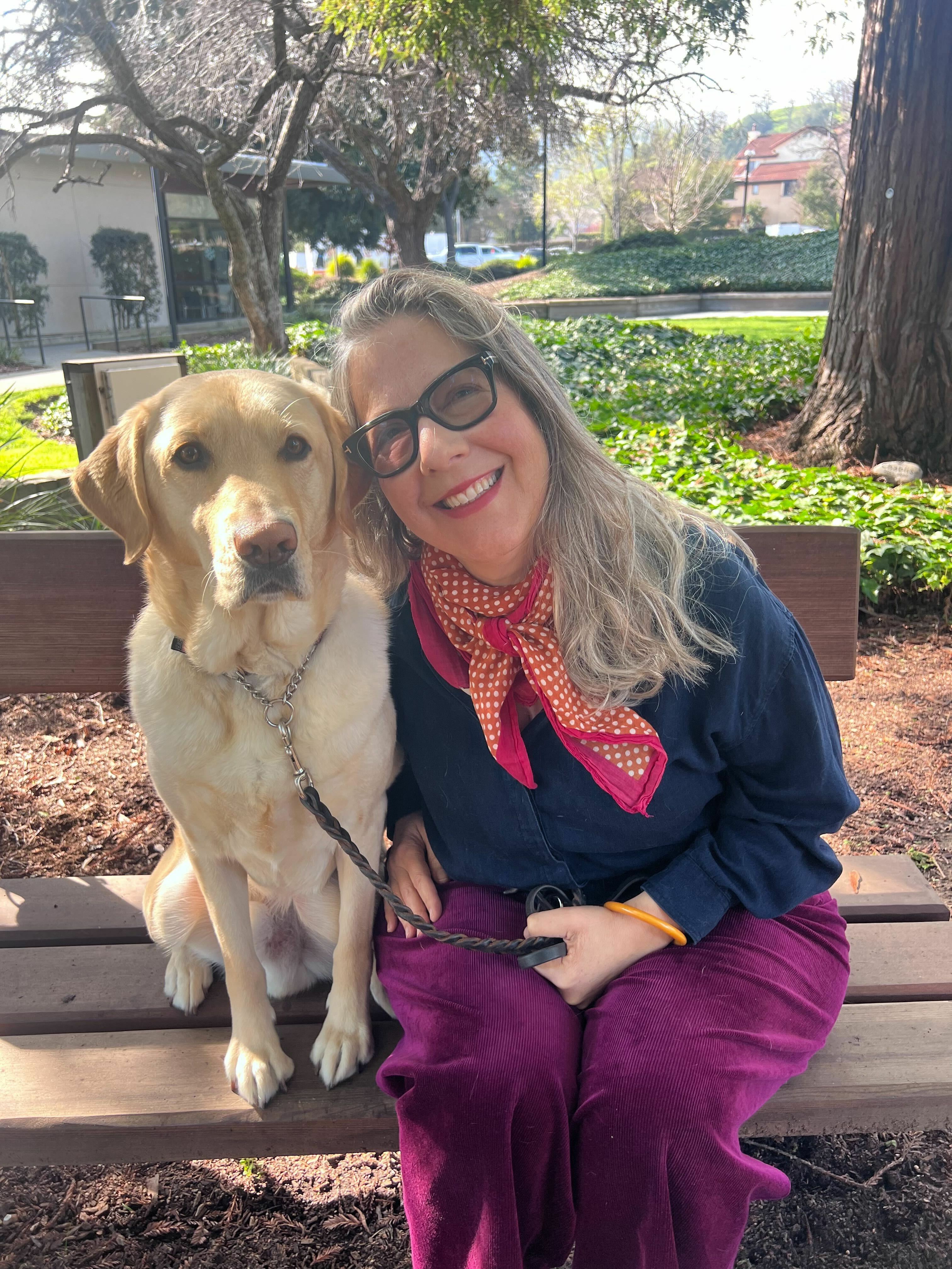 Lynn sits beside a yellow Lab on a bench under a shady tree. She has a brightly colored scarf around her neck and smiles at the camera while leaning her head into the dog's.