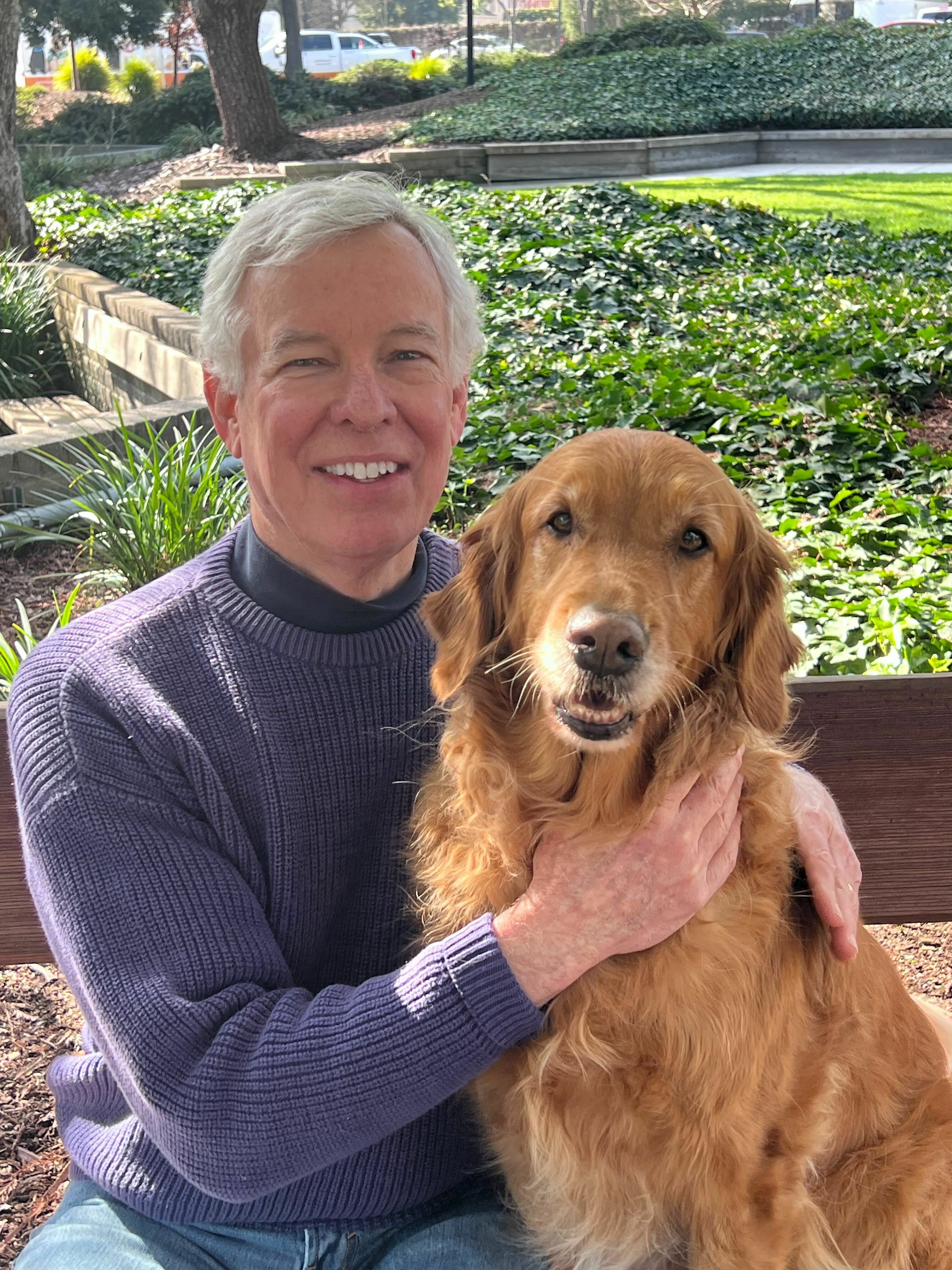 Chris sits on a bench under a shady tree with a Golden Retriever seated at his side. Both Chris and the dog smile widely at the camera.