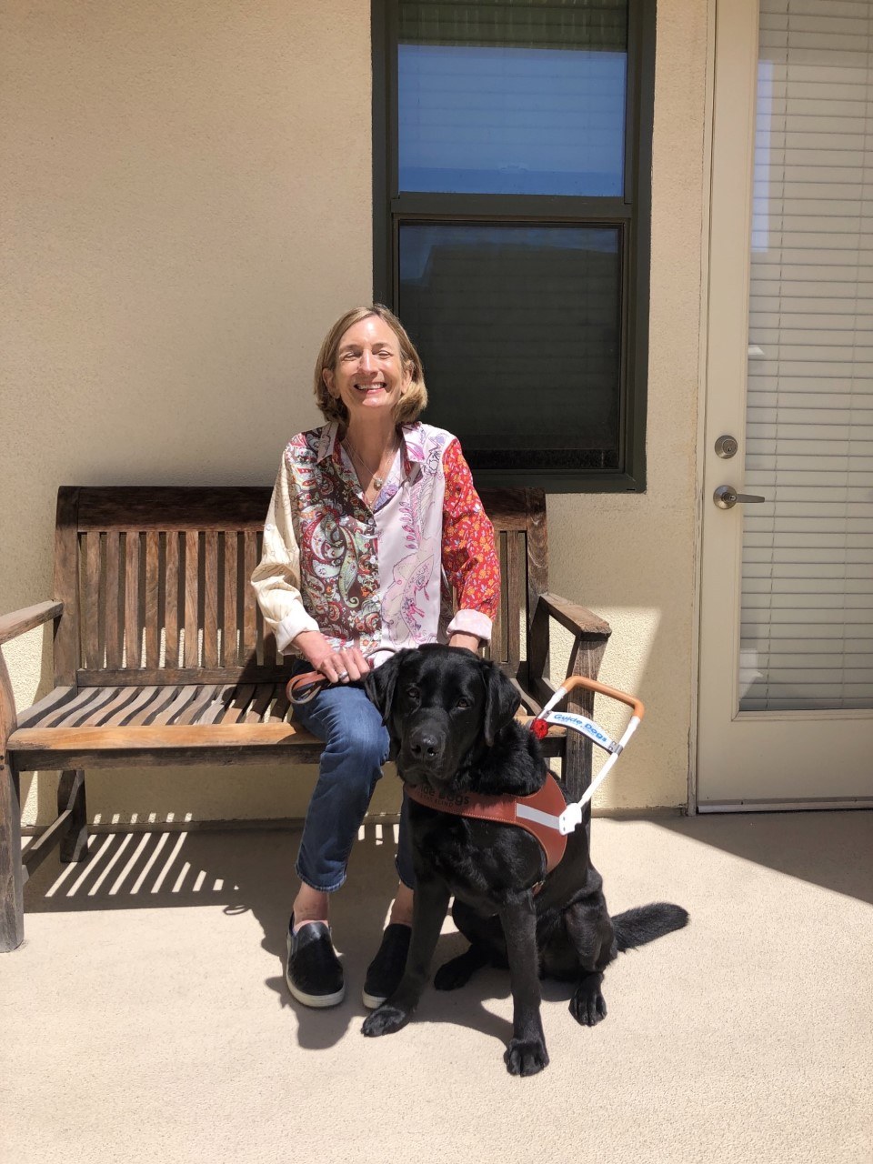 Kelly is seated on a sunny bench beside her black Lab guide dog.
