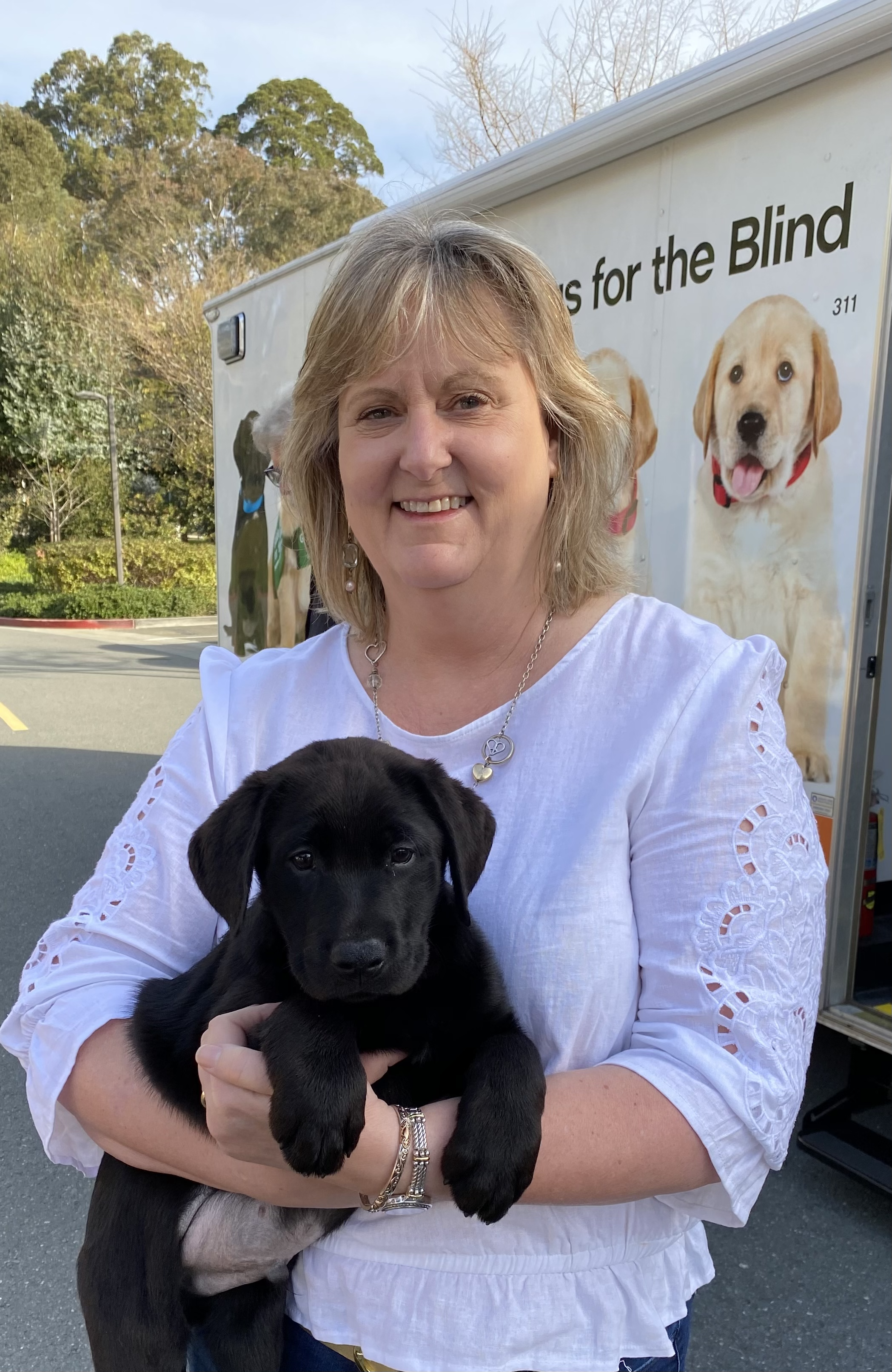 Sheri holds a black Lab puppy in front of the GDB Puppy Truck. She has blonde hair and is wearing a white top.