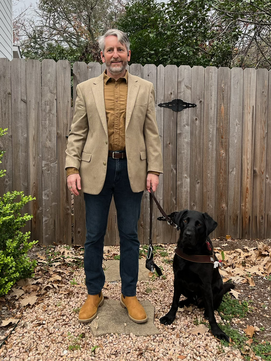 A man in a sport coat stands on a gravel path next to a black Lab guide dog with a wooden fence in the background.