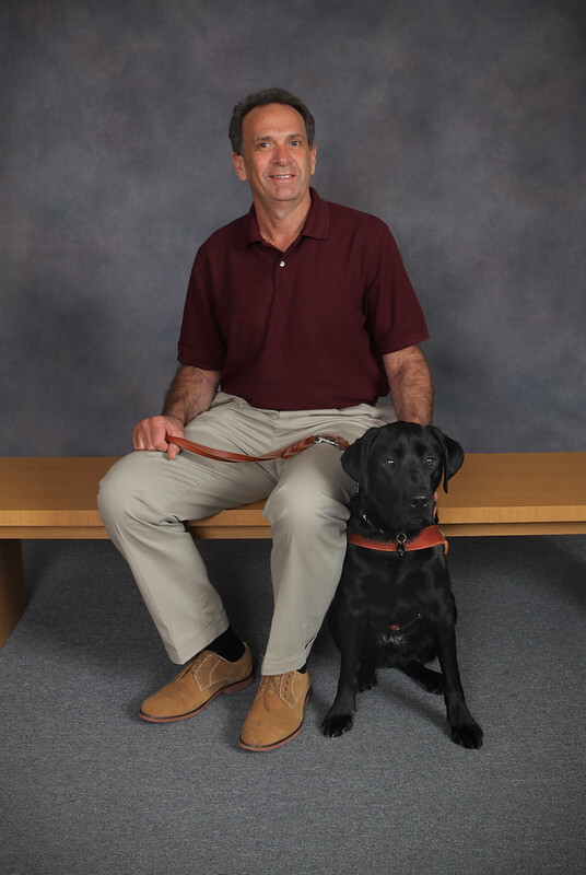 Posed portrait of a man sitting on a bench with a black Lab guide dog at his side.