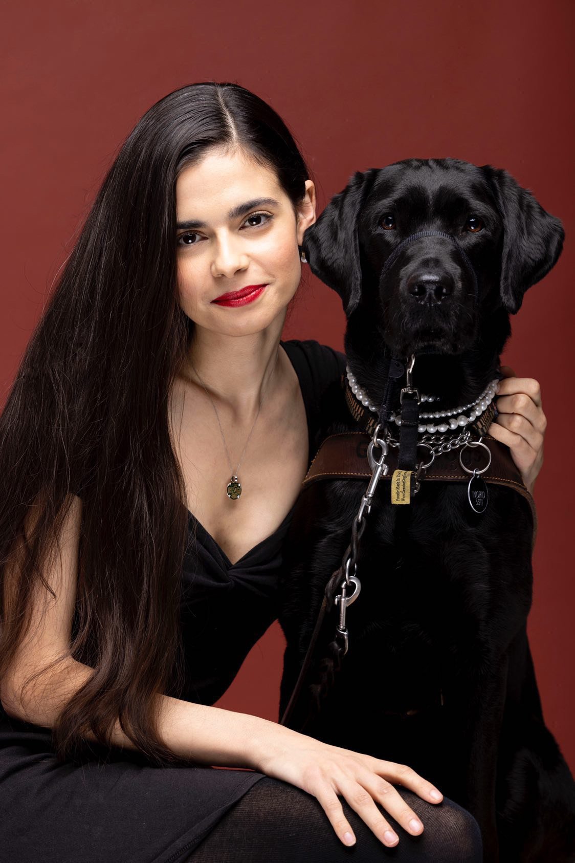 A woman with long black hair kneels against a rich brown backdrop. She has her arm around her black Lab guide dog. The guide dog is wearing her harness and a simple string of pearls with her collar.