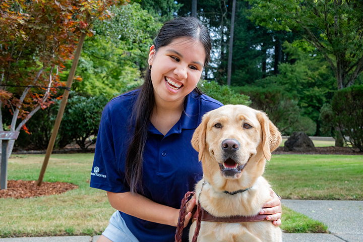 A smiling young woman kneels next to a smiling yellow Lab guide dog.