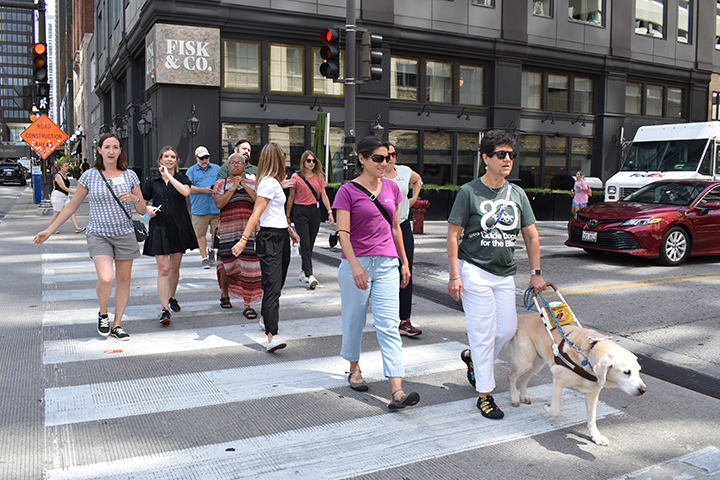 GDB client Pam Berman with guide dog Gumbo, and GDB Field Service Manager Lauren Ross, lead a group of GDB O&M Seminar participants across a busy street in downtown Chicago.