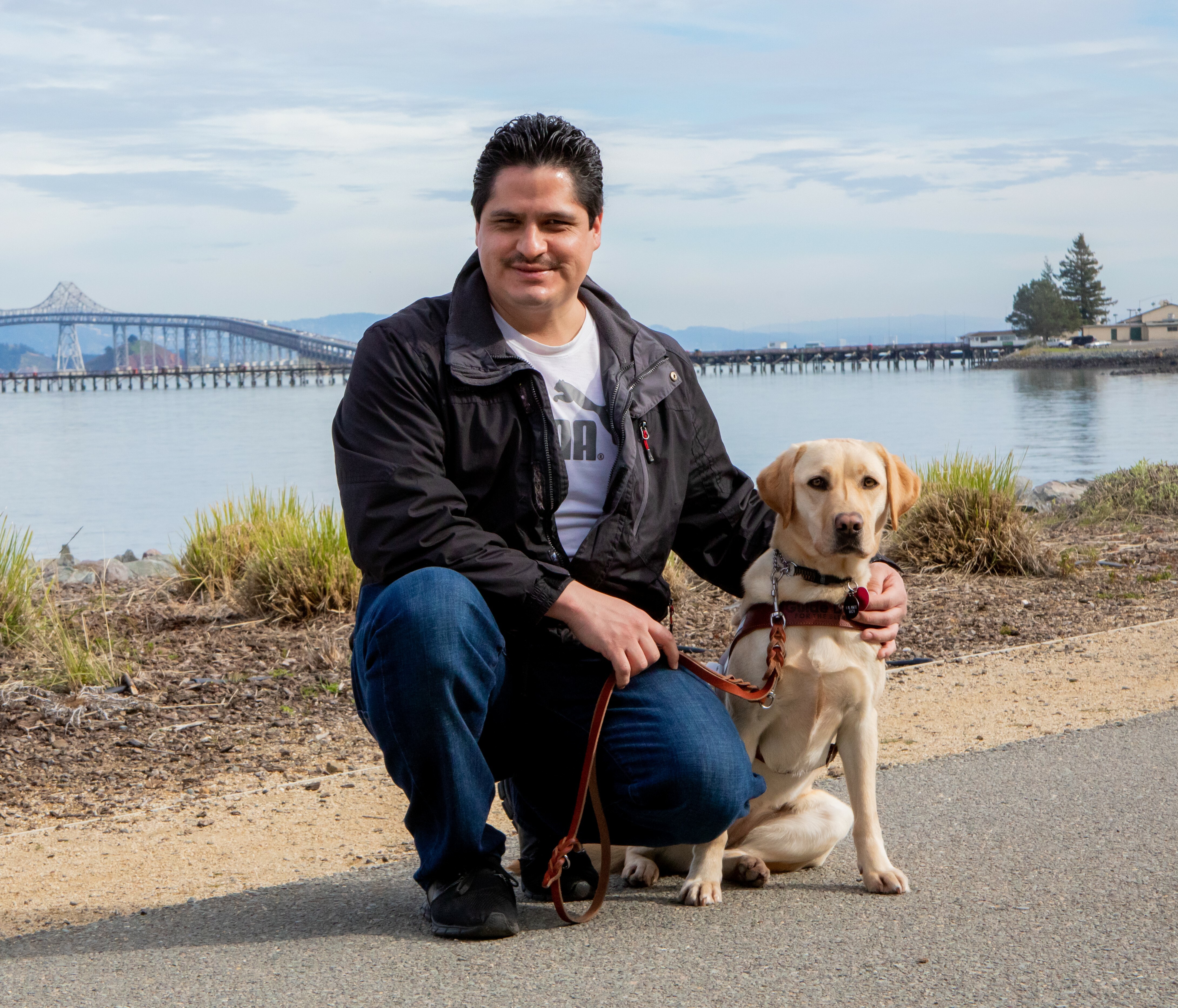 Ever Arreola kneels beside his yellow Lab guide dog, Falante, in front of the SF Bay.