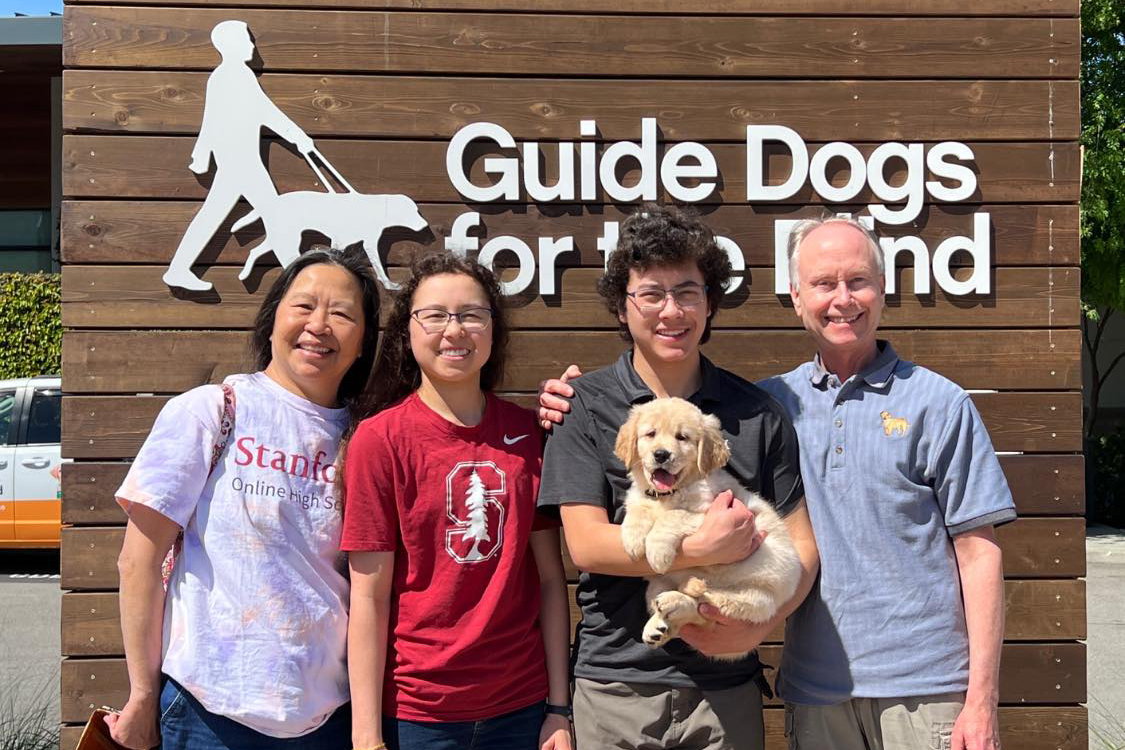 Larry Bowman (far right) poses with his wife and two children with guide dog puppy Sandra in front of a sign with Guide Dogs for the Blind's logo.