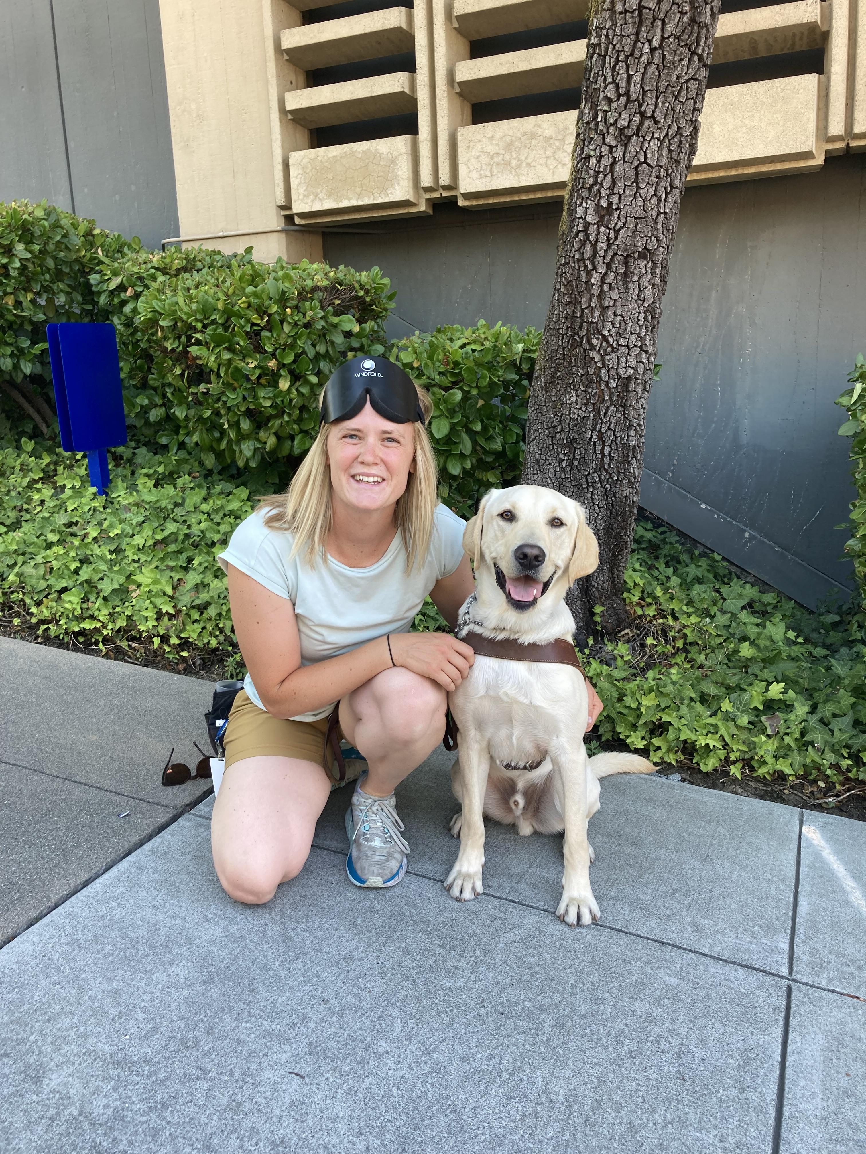 Jessica kneels beside a yellow Lab guide dog. She is wearing a blindfold flipped up on her head and both look smiling into the camera.