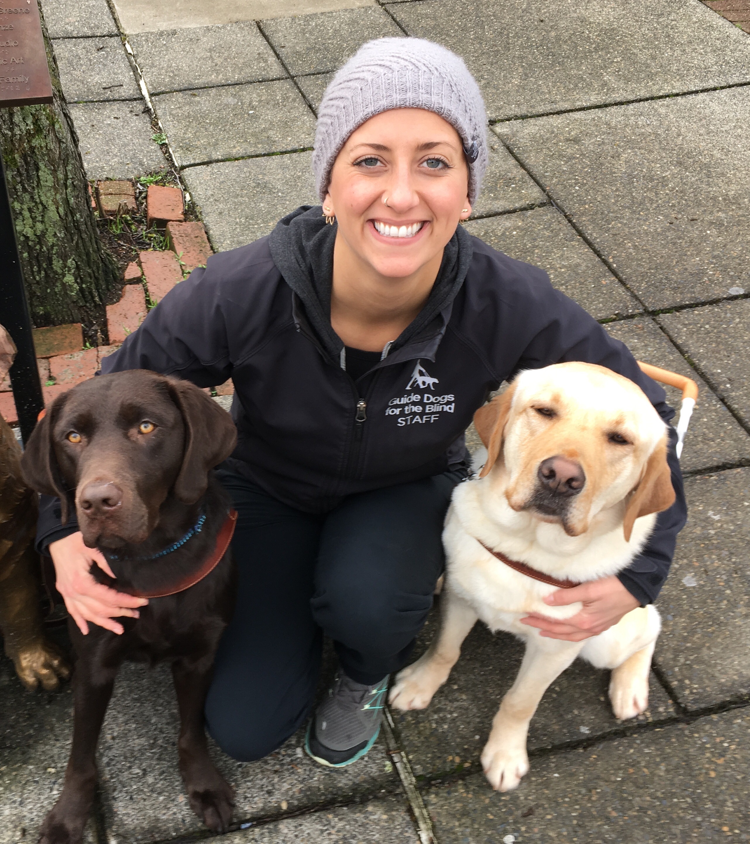 Guide Dog Mobility Instructor Megan Dodder kneeling with two guide dogs, a chocolate Lab and a yellow Lab.