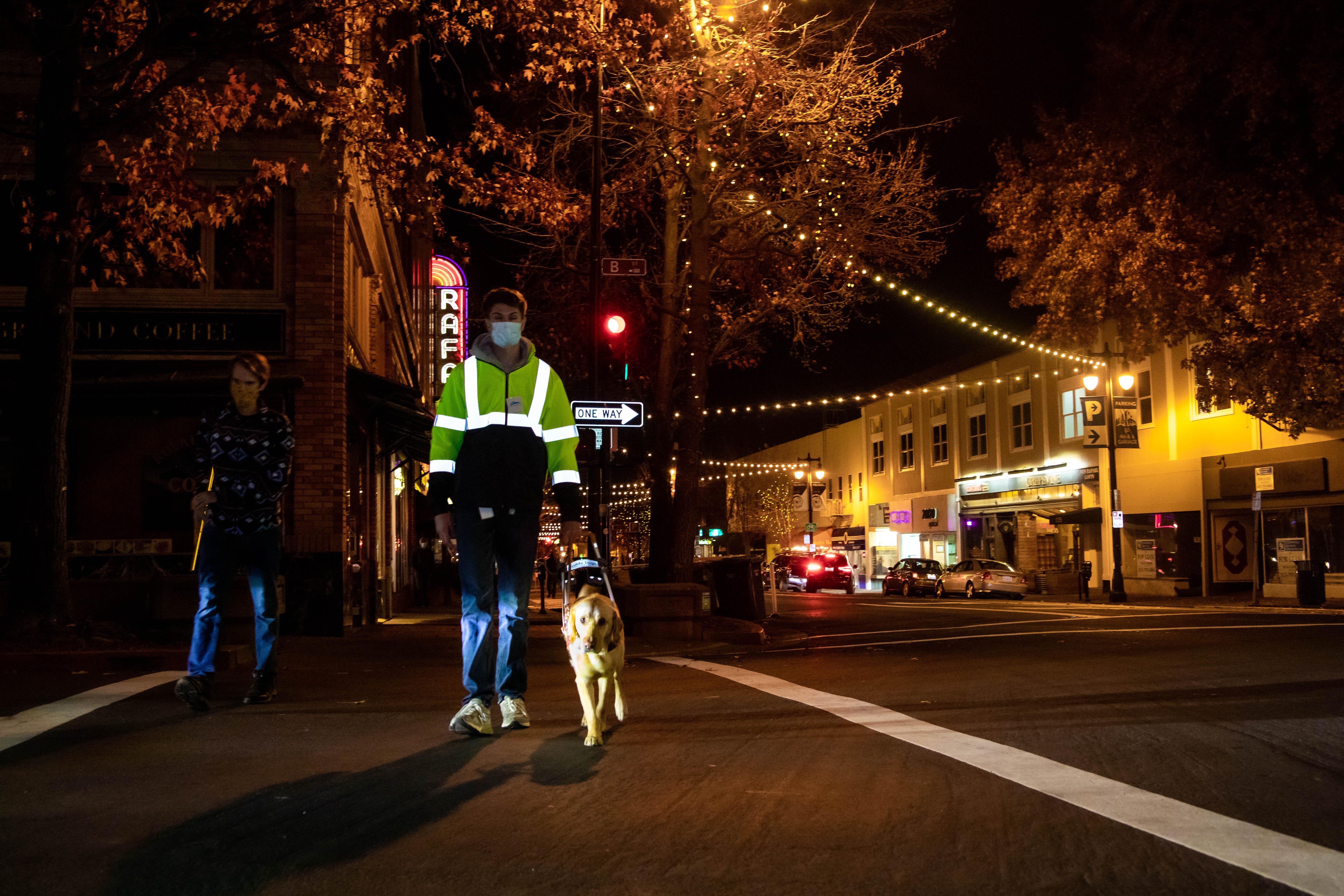 A young man wearing a reflective safety jacket walking with his guide dog across a street at night.