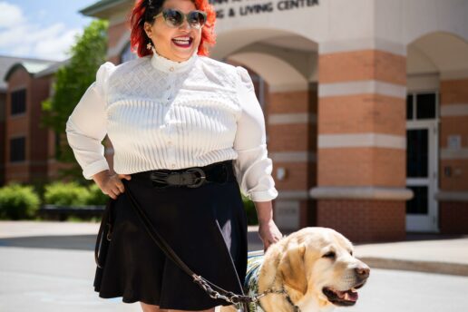 Melba stands with her yellow Lab guide dog Chad on the campus of the college where she is tenured professor. She  smiles widely at the camera.