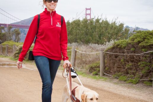 In this photo, Jane walks with her yellow Lab guide dog, Pilaf, along a coastal trail with the Golden Gate Bridge in the background. Jane smiles while looking forward and Pilaf looks focused on their destination.