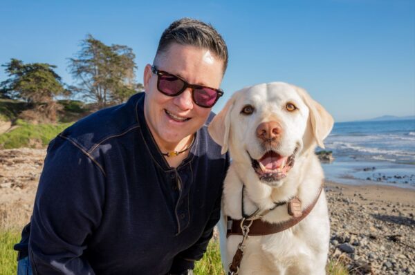 A person sits on a beach with their yellow Lab guide dog.
