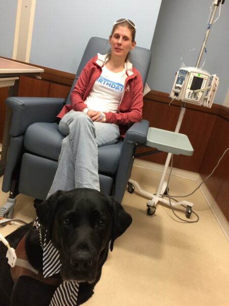 A woman sits in the hospital with her black Lab guide dog at her feet.