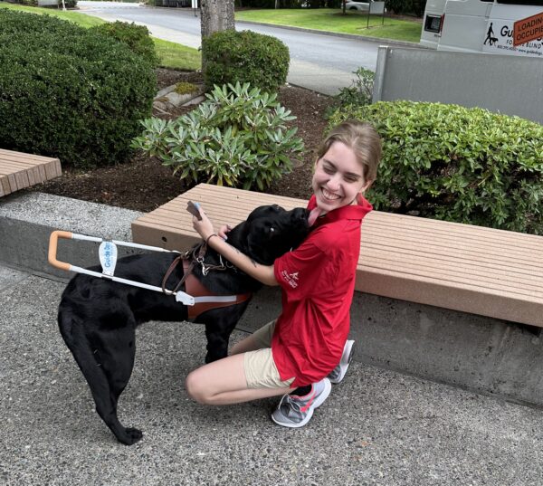 A woman kneels with her arms around a black Lab guide dog in harness.