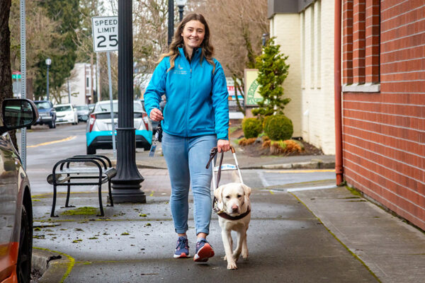 A woman walks with a guide dog on a city sidewalk.