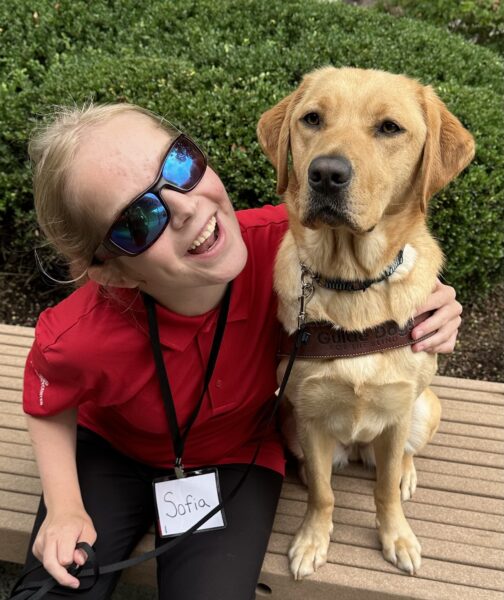 A woman smiles with her arm a yellow Lab guide dog.