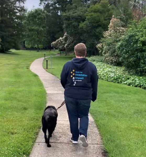 A woman wearing the PAWSitive Impact sweatshirt walks on a campus path with her black Lab dog.