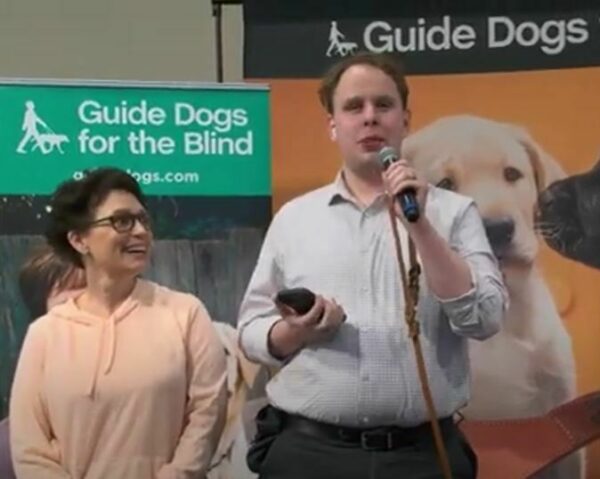 Derek Dittmar speaks into a microphone during a Graduation ceremony on our California campus. Volunteer Puppy Raiser Annie Martin stands next to him. Colorful GDB banners are in the background.