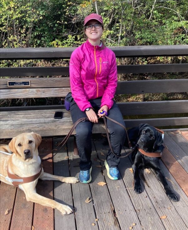 A woman sits on a bench with two guide dogs lying at her feet.