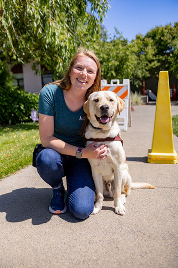 A smiling woman kneels next to a smiling guide dog on a sidewalk with traffic cone obstacles in the background.