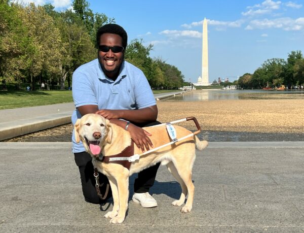 Brian kneels behind his yellow Lab guide dog with the Washington Monument in the background