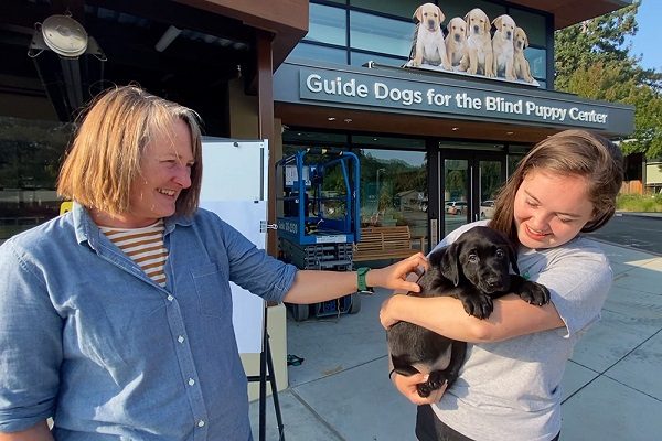 Two volunteer puppy raisers embrace a black Lab puppy in front of the GDB puppy center.
