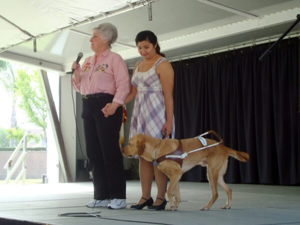 Two women stand on a GDB graduation stage with a guide dog and speak.