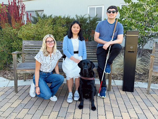 Three young adults pose for a photo; one woman kneels in front of a bench, a second woman sits on the bench with a black Lab guide dog at her feet, and a man sits on the arm of the bench and holds a white cane.
