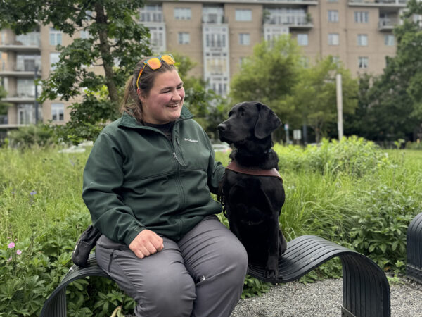 A woman sits in a bench next to a black Lab guide dog.