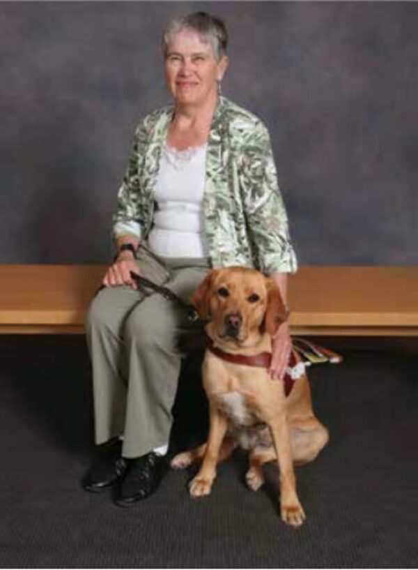 Judy sits on a bench with her around around  her yellow Lab guide dog Rosie.
