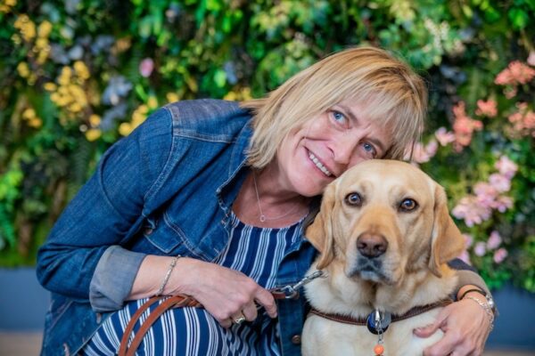 Woman embraces her yellow Lab guide dog in front of a colorful wall of flowers.