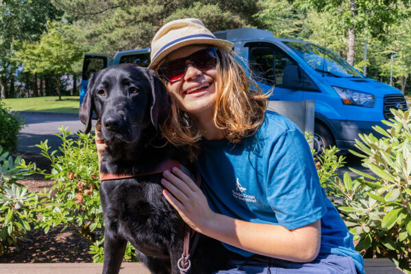 A teen boy with long blond hair smiles and hugs a black Lab guide dog.