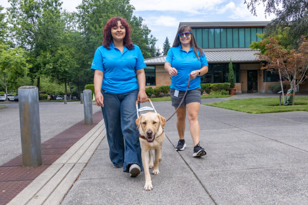 Two smiling women walk with a yellow Lab guide dog.