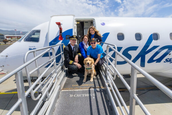 An airline pilot and flight attendant pose for a photo in front of an open door to an airplane with two people and a guide dog puppy.