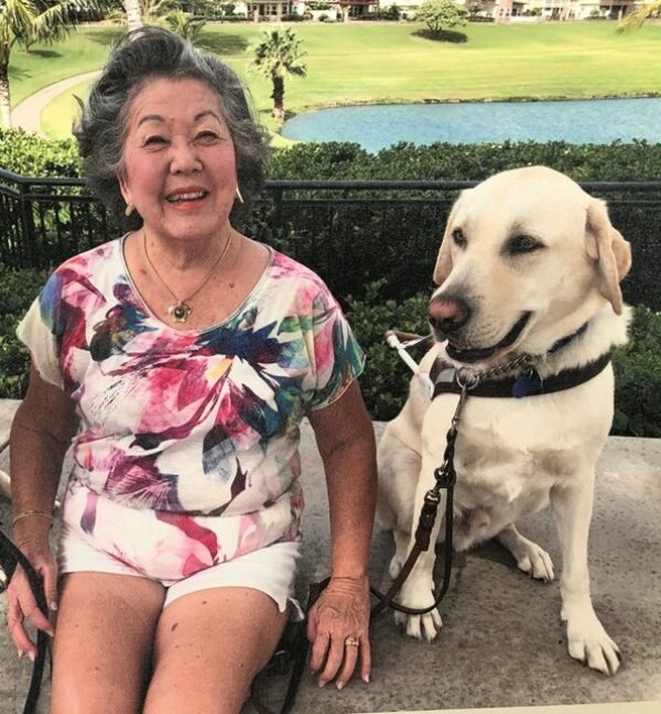 A woman sits next to her yellow Lab guide dog in front of a body of water, grass, and trees.
