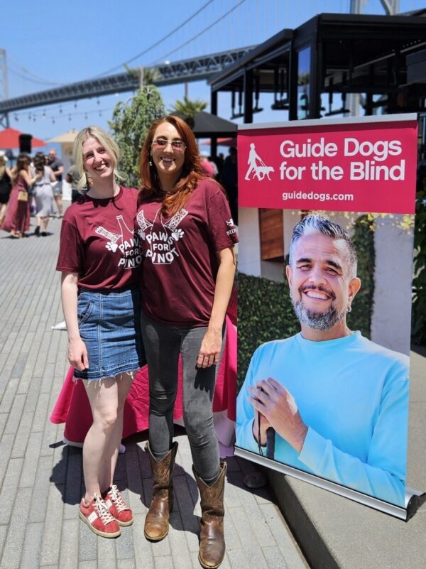 Two women stand at an outdoor event near a GDB banner. A bridge appears in the background against a blue sky.