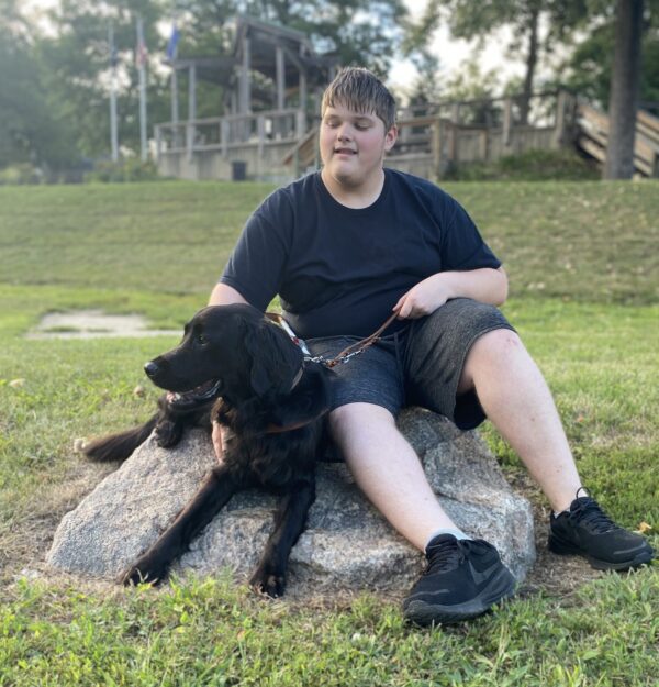 A teenage boy sits on a rock in a grassy field with his Golden Retriever black Lab cross guide dog.