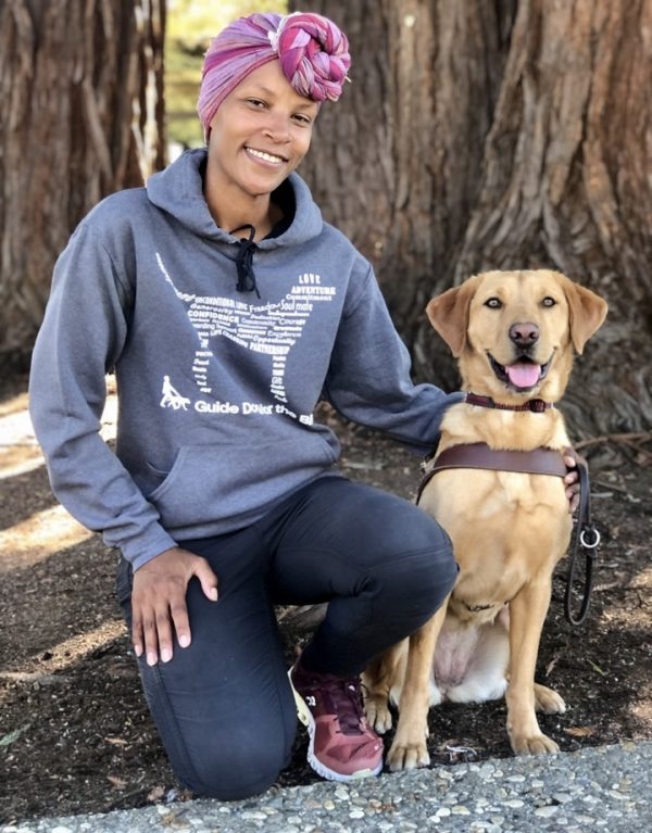 A GDB staff member kneels beside a yellow Lab guide dog. She is wearing a gray hoodie with the outline of a dog on the front.