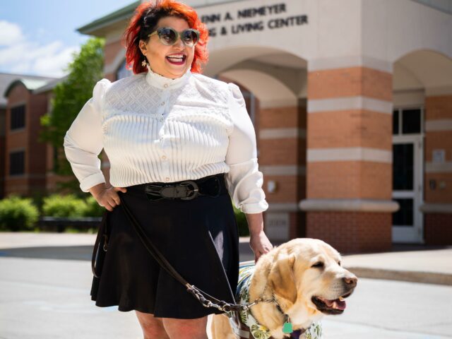 Melba stands with her yellow Lab guide dog Chad on the campus of the college where she is tenured professor. She smiles widely at the camera.