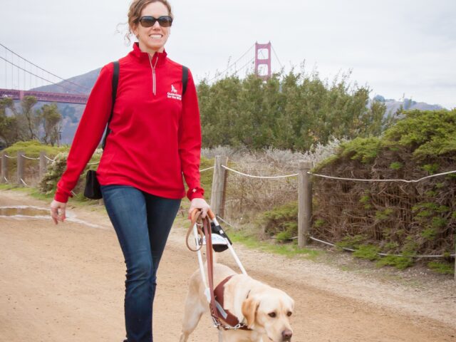 In this photo, Jane walks with her yellow Lab guide dog, Pilaf, along a coastal trail with the Golden Gate Bridge in the background. Jane smiles while looking forward and Pilaf looks focused on their destination.