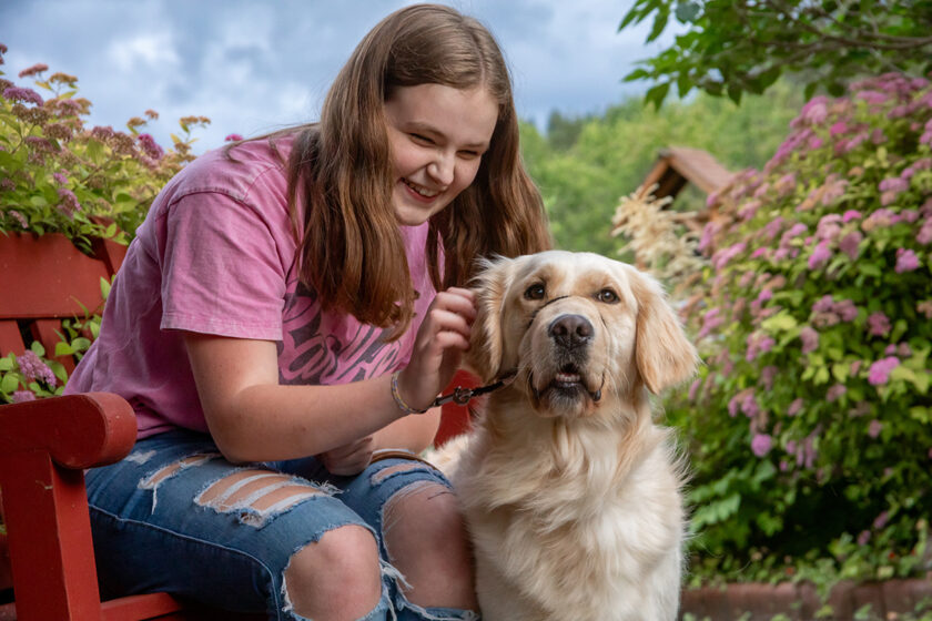 A girl sits on a bench in a garden and smiles at a Golden Retriever guide dog. There are pink flowers in the background that match her pink shirt.