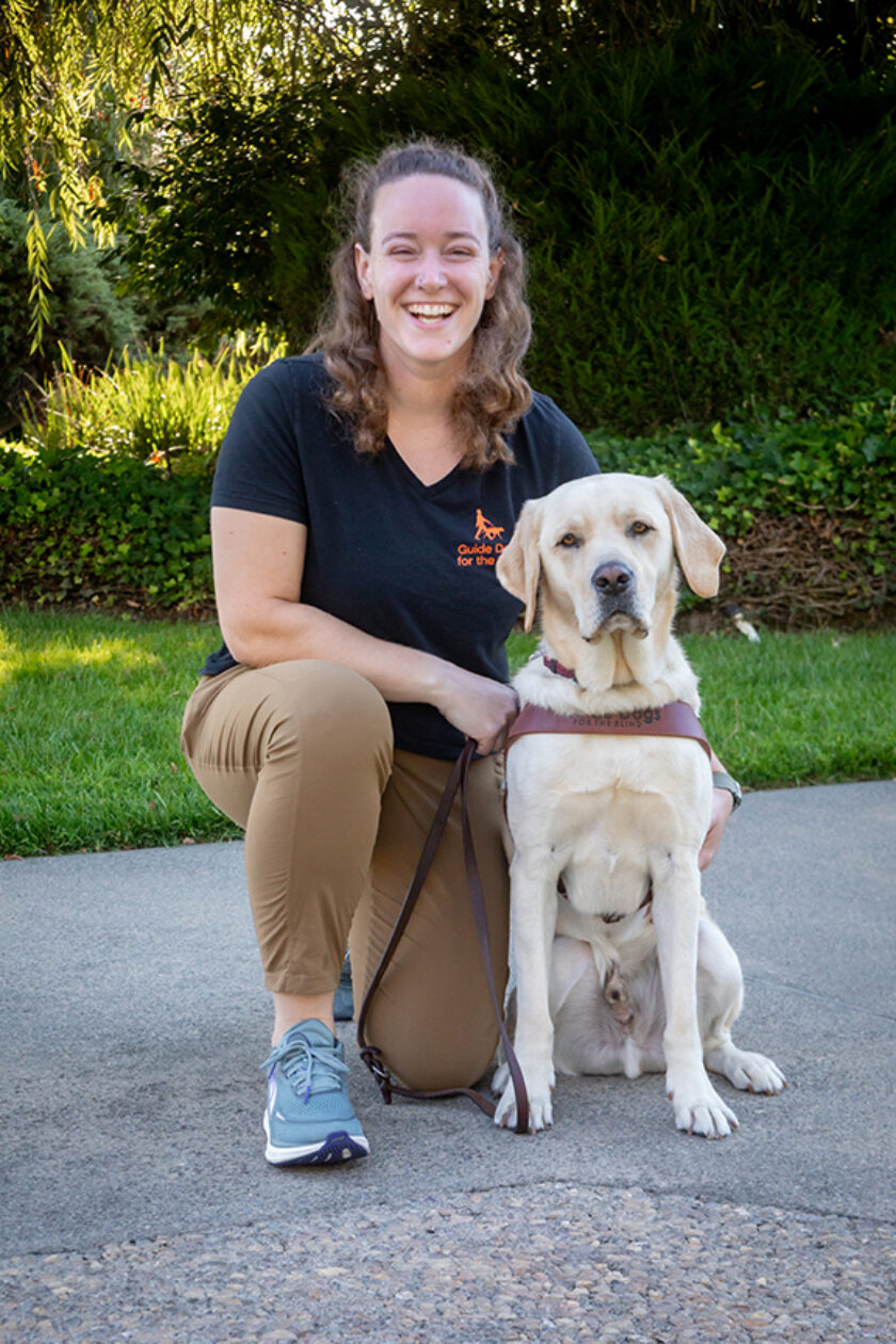 A woman kneels next to a yellow Lab guide dog.