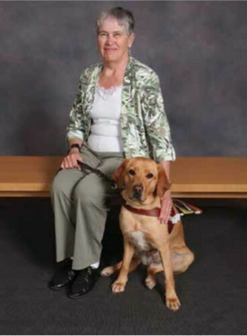 Judy sits on a bench with her around around her yellow Lab guide dog Rosie.